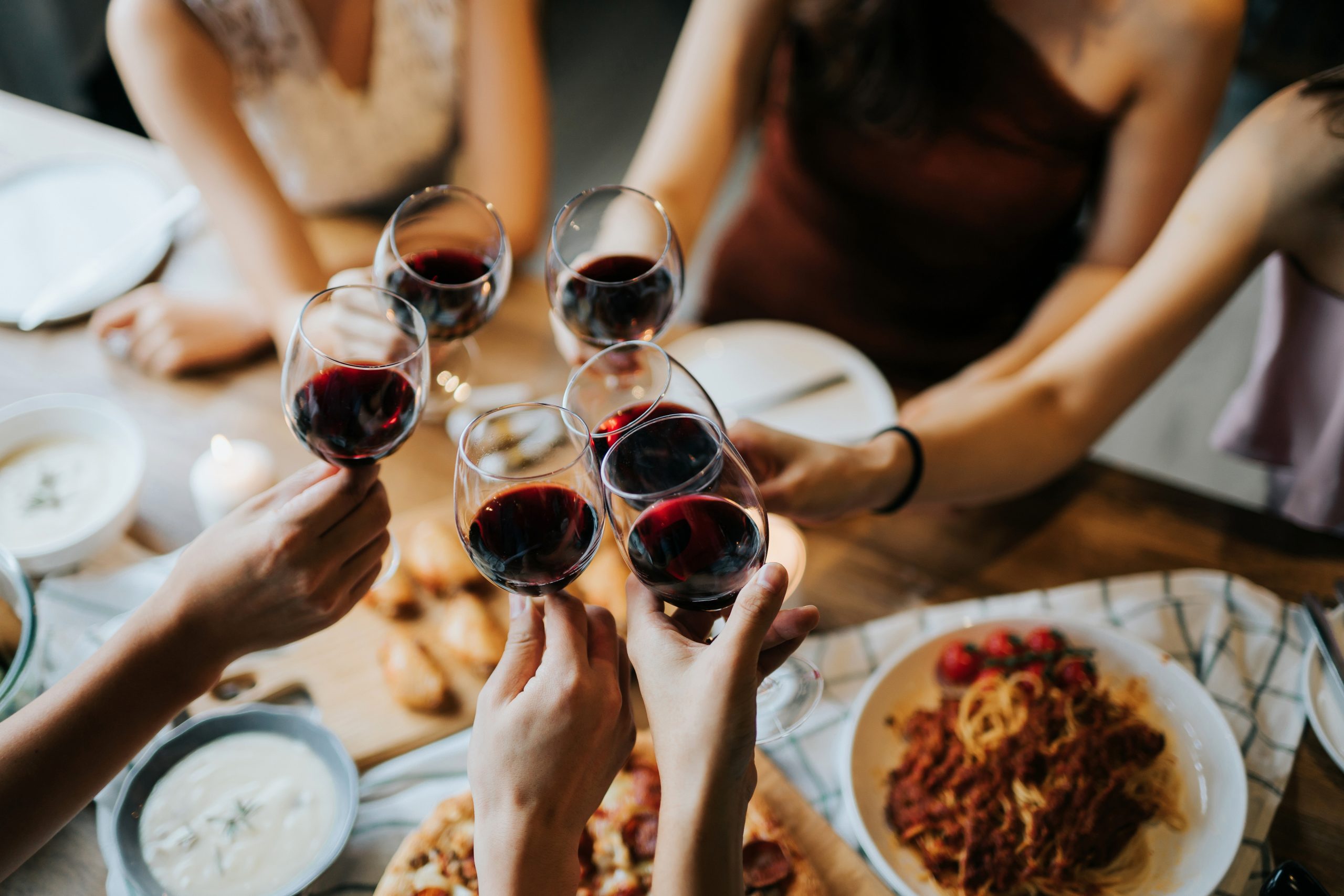 Hände stoßen mit Rotweingläsern über einem Tisch mit Pasta an – Symbolbild für typische Auslöser einer Histaminintoleranz
